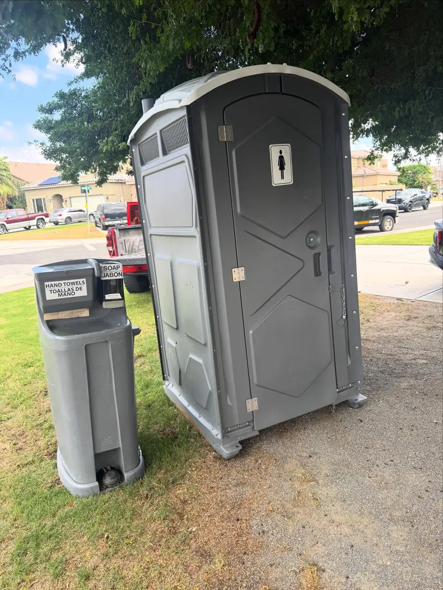 Gray porta potty beside a 2-sided hand wash station on a lawn with homes behind